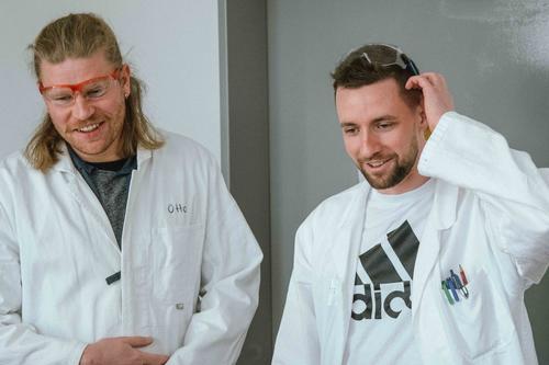 Zwei Männer in weißen Laborkitteln und Schutzbrillen stehen nebeneinander und lächeln. Der linke trägt ein Namensschild mit „Otto“, der rechte hat bunte Stifte in der Brusttasche. Beide wirken entspannt und freundlich.