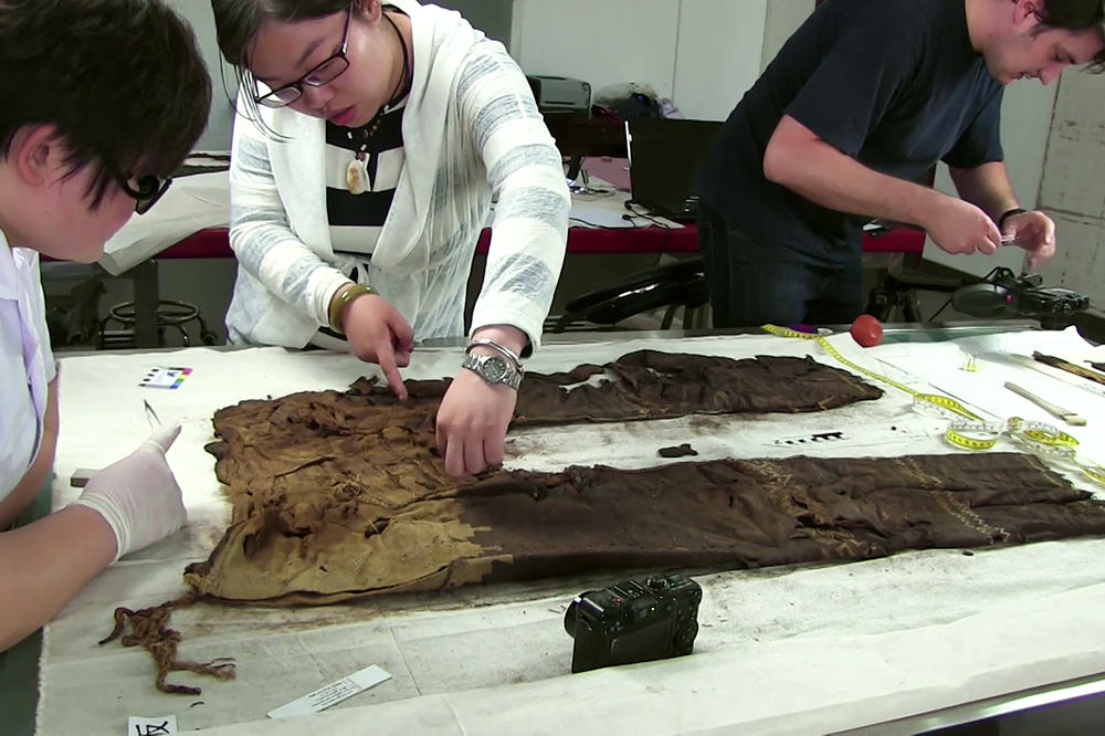 Wissenschaftlerinnen und WIssenschaftler in weißen Laborkitteln und Gummihandschuhen untersuchen auf einem Tisch eine jahrtausende alte Hose.