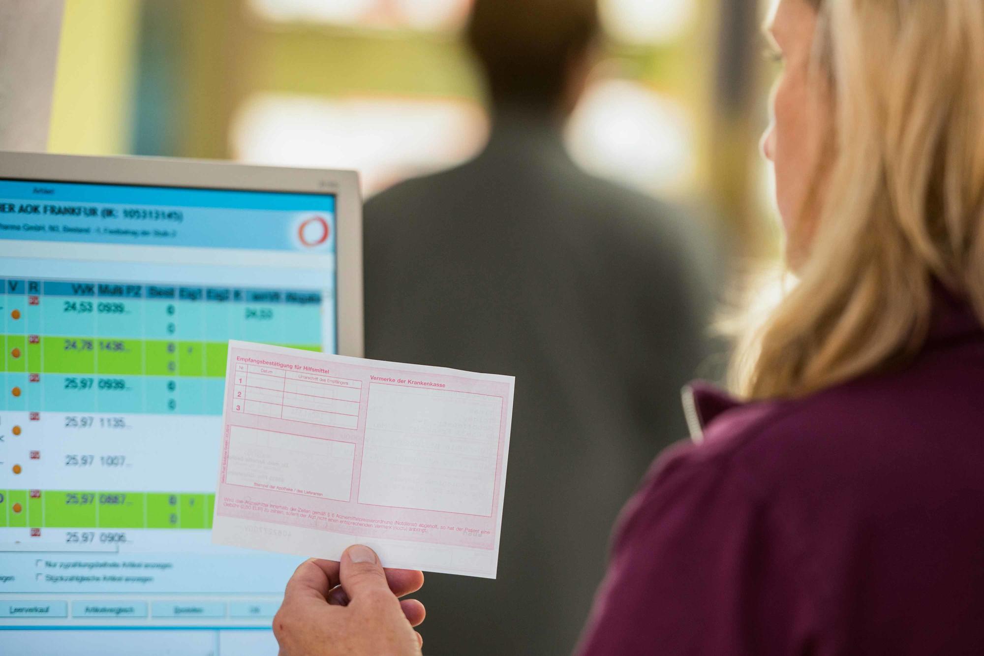 Rezept für Arzneimittel, das eine Apothekerin vor einem Monitor hoch hält